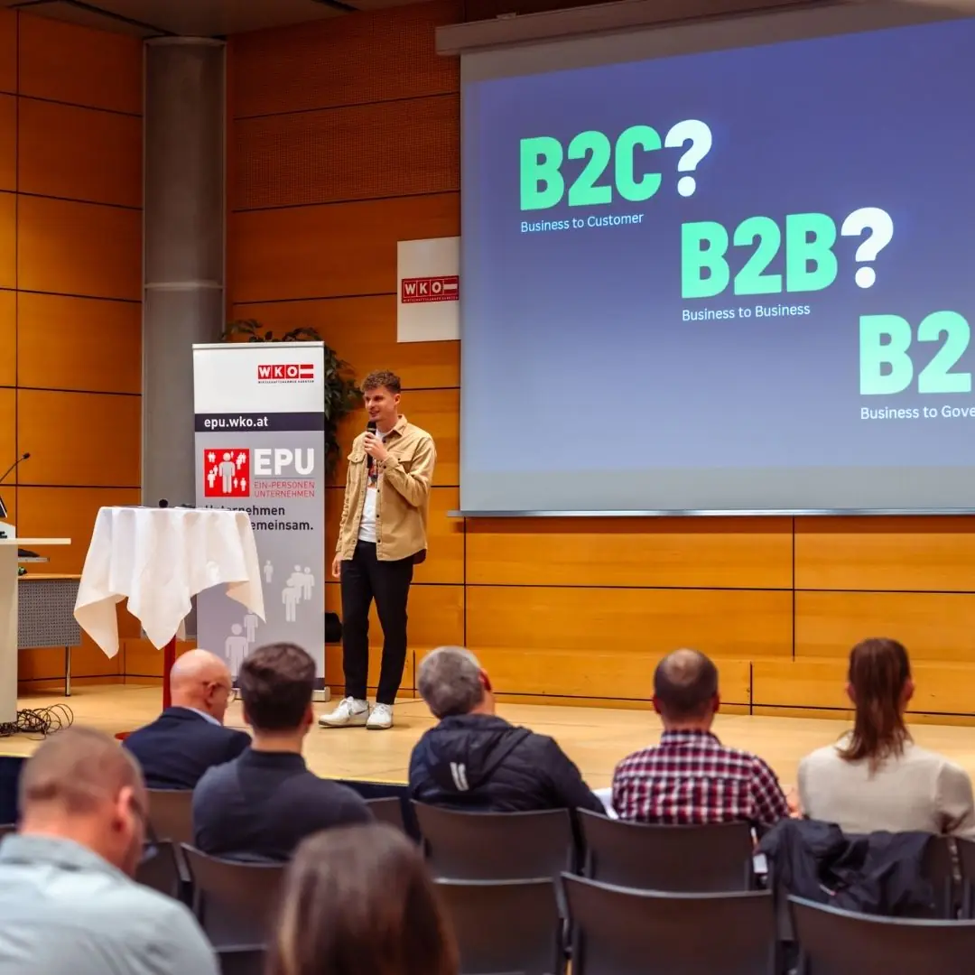 Florian Semmler auf der Bühne mit Mikrofon, Folie zu B2C/B2B, WKO-EPU-Roll-up und Publikum im Vordergrund
