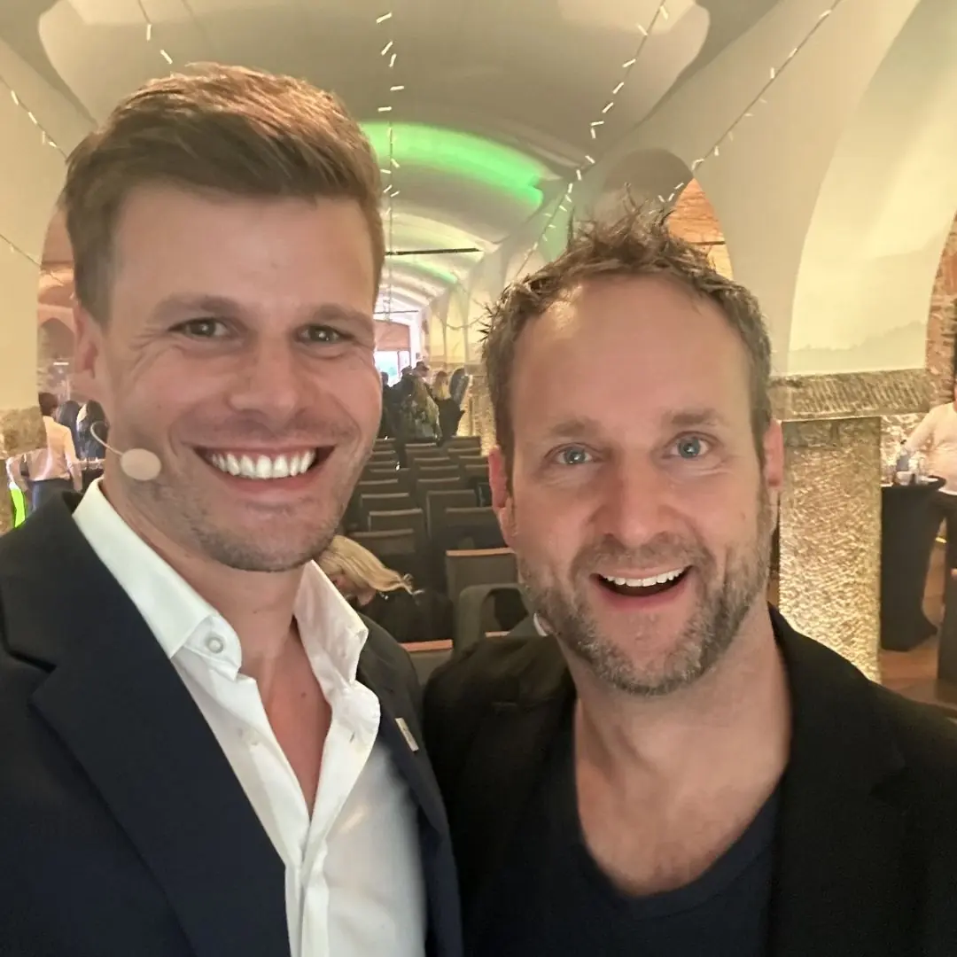 Florian Semmler mit Headset neben Co-Moderator, Selfie im Veranstaltungssaal mit Bogendecke und Bestuhlung