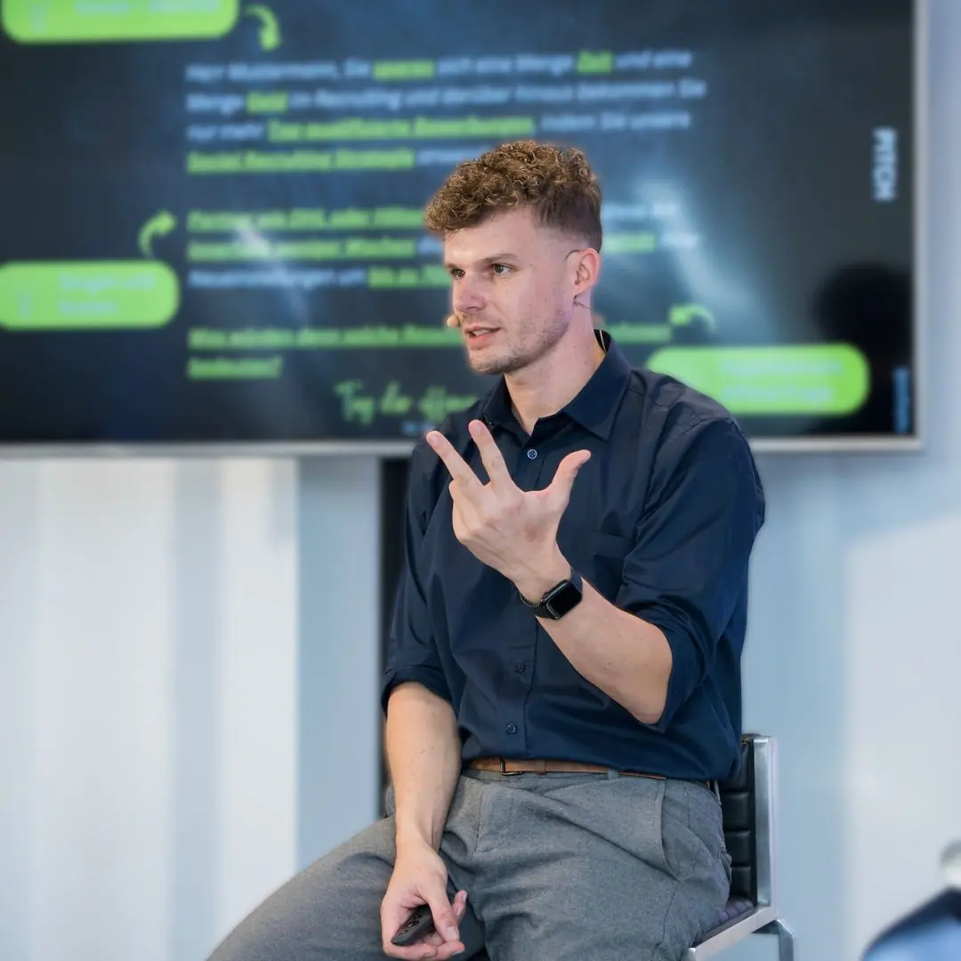 Workshop oder Vortrag: Referent mit Headset auf Hocker vor großem Screen mit Pitch-Folie und grünen Grafiken, Klicker in der Hand